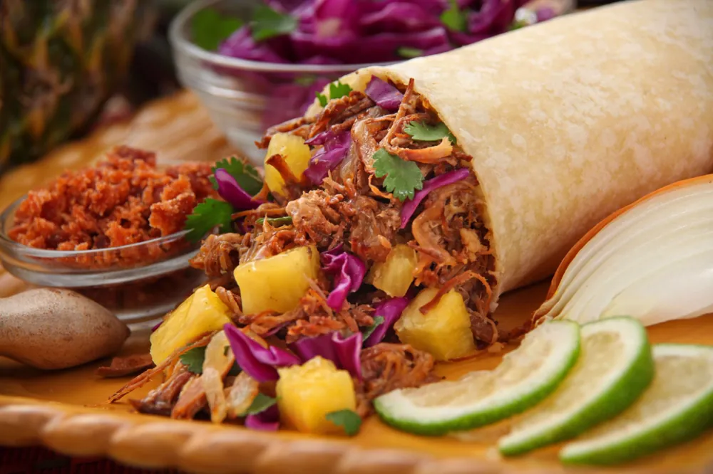 A burrito opened to show shredded meat, pineapple, red cabbage and cilantro, with lime slices