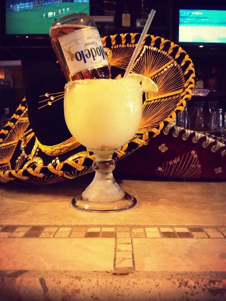 A frozen margarita with salted rim and lime on the bar, in front of a gold-embroidered sombrero